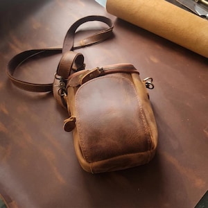 May include: A brown leather crossbody bag featuring a vertical rectangular shape, a front flap pocket with a zipper closure, and an adjustable shoulder strap attached with metal hardware. The bag has a smooth, matte finish with visible stitching.