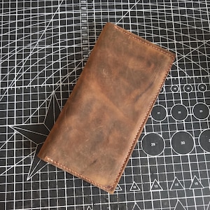 May include: A rectangular brown leather wallet featuring a distressed finish and visible stitching along the edges. The wallet is positioned diagonally on a black cutting mat marked with white grid lines, circular patterns, and numerical indicators.