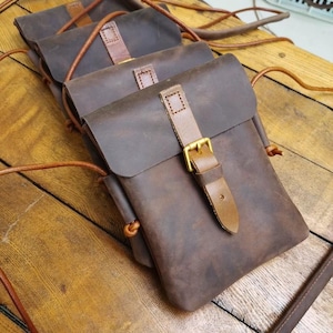 Pode incluir: Uma série de bolsas de couro castanho escuro dispostas em fila. Cada bolsa apresenta uma aba dobrável fixada por uma tira de couro castanho mais claro com uma fivela de metal cor de latão. Cordões de couro finos servem como alças.