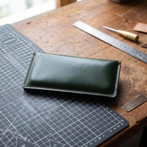 Puede incluir: Una cartera rectangular de cuero verde oscuro con costuras visibles a lo largo de los bordes. La cartera está colocada sobre una alfombrilla de corte negra junto a una regla de metal y una pequeña herramienta de latón, con una placa de metal con la inscripción Crafted in Hands.