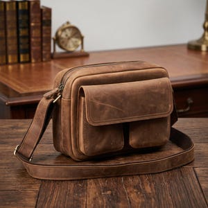 Puede incluir: Un bolso bandolera de cuero marrón con forma rectangular, que cuenta con un compartimento principal con cremallera y dos bolsillos delanteros con cierre de solapa. Incluye una correa ajustable y tiene un acabado mate y texturizado.