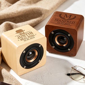 May include: Two square wooden speakers with circular black drivers. The light wood speaker is engraved with a hat, bowtie, and the text JUSTIN BEST MAN 02.05.2025. The dark wood speaker is engraved with a laurel wreath, the letter M, MANFRED, and 02.05.2025.