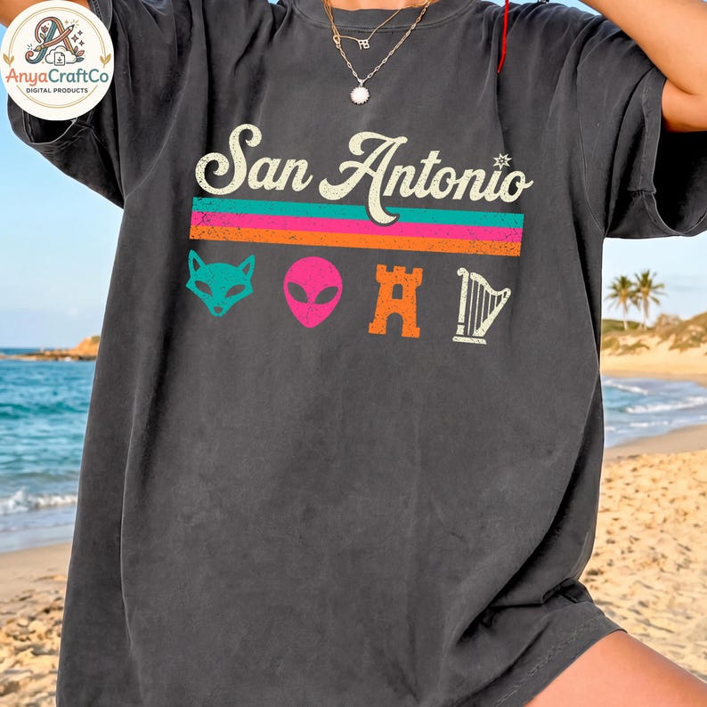 May include: Dark gray t-shirt featuring the text "San Antonio" in a vintage style. Below the text are colorful graphics including a fox, alien, castle, and harp. The shirt is a relaxed fit.