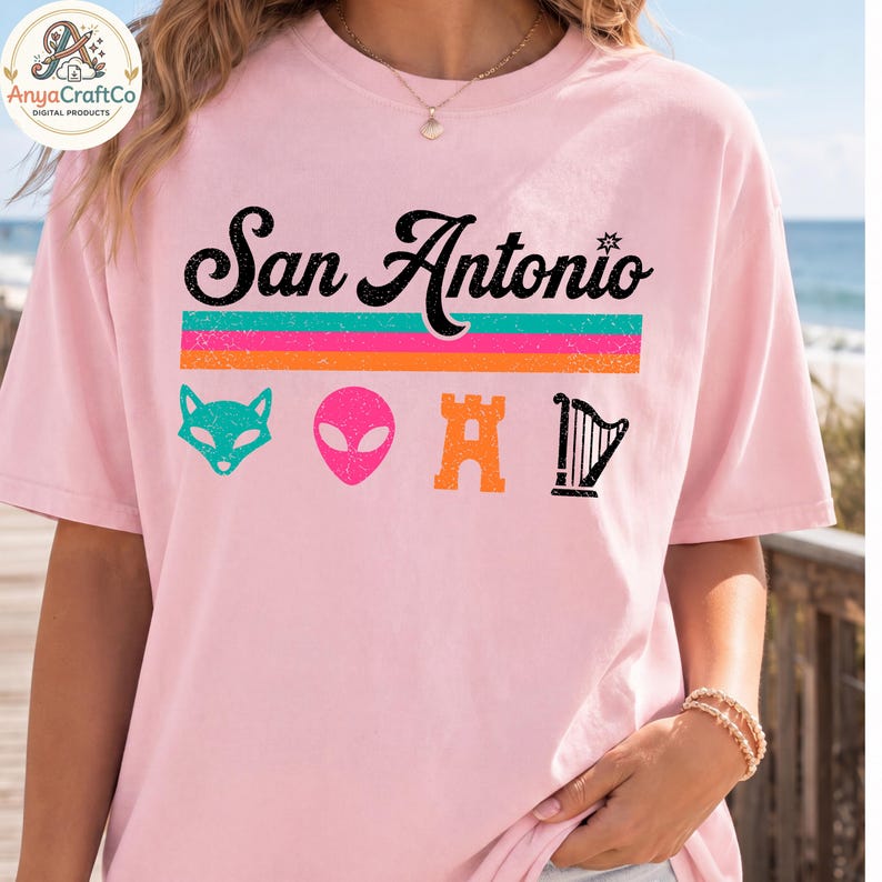 May include: A light pink t-shirt with the words "San Antonio" in black script above a horizontal stripe design in teal, orange, and pink. Below the stripes are teal fox, pink alien, orange castle, and black harp graphics. The shirt is a casual style.