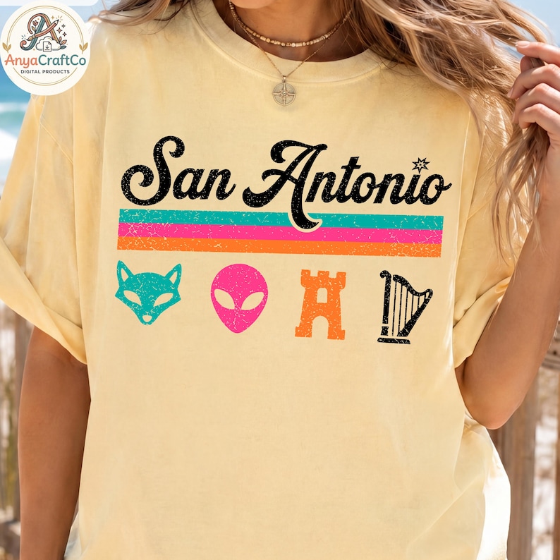 May include: A light yellow t-shirt with the text "San Antonio" in black script above a horizontal striped design in teal, pink, and orange. Below are cartoon graphics of a fox, alien, castle, and harp.