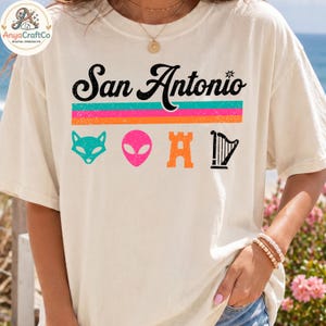 May include: Cream-colored t-shirt featuring the text "San Antonio" in black, above a horizontal stripe design in teal, pink, and orange. Below the text are teal fox, pink alien, orange castle, and black harp graphics. The shirt is worn by a person.
