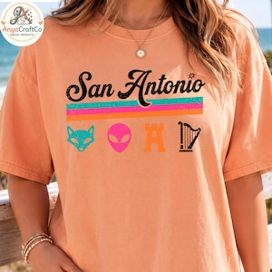 May include: Peach-colored t-shirt featuring the text "San Antonio" in black script above a horizontal stripe design in teal, pink, and orange. Below the text are four colorful icons: a teal fox, a pink alien, an orange castle, and a black harp.