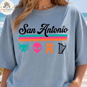 May include: A light blue t-shirt with the text "San Antonio" in black script above a horizontal stripe design in pink, orange, and green. Below the text are four colorful graphic icons: a fox, an alien, a castle, and a harp.