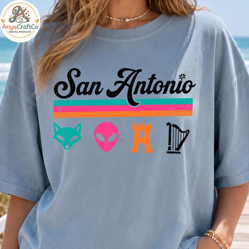 May include: A light blue t-shirt with the text "San Antonio" in black script above a horizontal stripe design in pink, orange, and green. Below the text are four colorful graphic icons: a fox, an alien, a castle, and a harp.