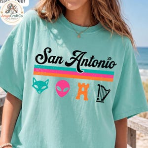 May include: A light teal t-shirt with the text "San Antonio" in black script above a horizontal stripe design in pink, orange, and teal. Below the text are cartoon graphics of a fox, alien, castle, and harp.