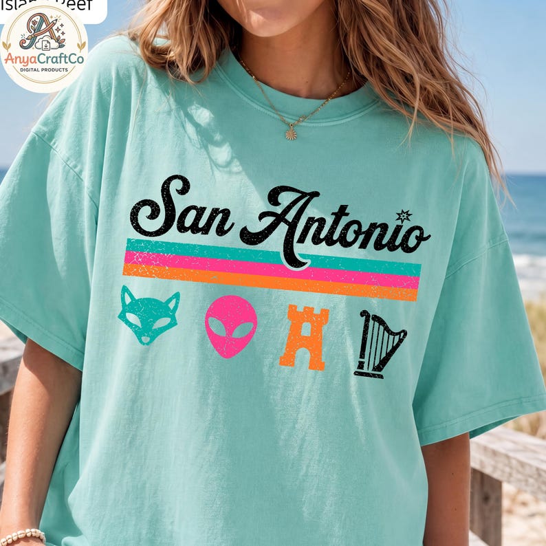 May include: A light teal t-shirt with the text "San Antonio" in black script above a horizontal stripe design in pink, orange, and teal. Below the text are cartoon graphics of a fox, alien, castle, and harp.