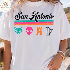 May include: White t-shirt with the text "San Antonio" in black script above a horizontal stripe design in teal, pink, and orange. Below the text are four colorful icons: a teal fox, a pink alien head, an orange castle, and a black harp.