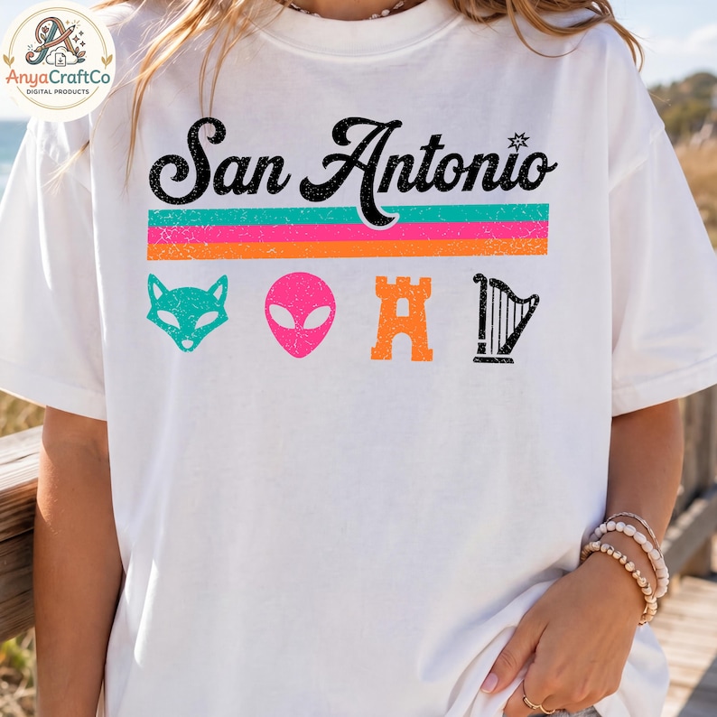 May include: White t-shirt with the text "San Antonio" in black script above a horizontal stripe design in teal, pink, and orange. Below the text are four colorful icons: a teal fox, a pink alien head, an orange castle, and a black harp.