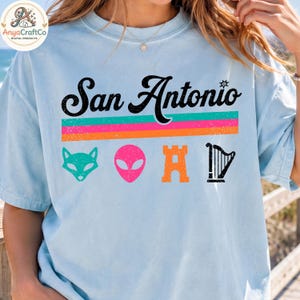 May include: Light blue t-shirt with the text "San Antonio" in black script above colorful horizontal stripes. Below are teal fox, pink alien, orange castle, and black harp graphics. The shirt is a casual style.