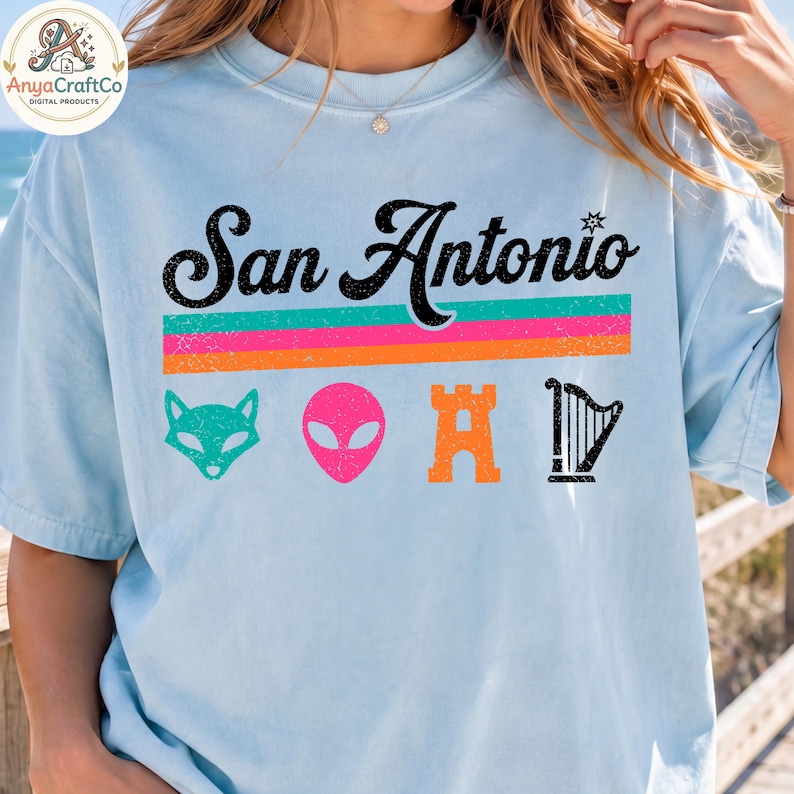 May include: Light blue t-shirt with the text "San Antonio" in black script above colorful horizontal stripes. Below are teal fox, pink alien, orange castle, and black harp graphics. The shirt is a casual style.