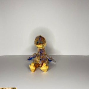 Puede incluir: Un pato de peluche con pico y patas amarillas, alas azules y cuerpo multicolor. El pato está sentado sobre una superficie blanca. El juguete es un Beanie Baby.