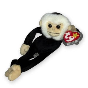 May include: A plush monkey toy with a black body, beige hands and feet, and a white fur face. The toy features a heart-shaped red and yellow Ty Beanie Original tag attached to its right side, displaying the text ty in white script.