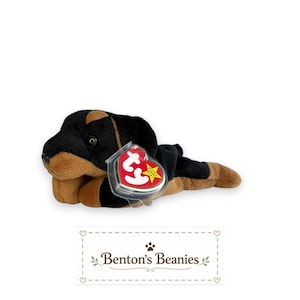 May include: A plush dog toy with black and tan fur, featuring a small black nose and dark eyes. A red heart-shaped tag with the white letters ty is attached to the side in a clear plastic protector. Below the toy is the text Benton's Beanies.