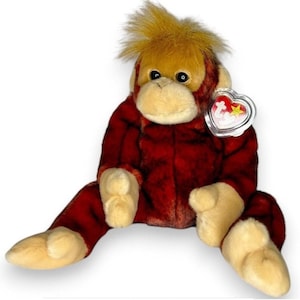 May include: A plush orangutan toy with dark red fur, a light tan face, and a tuft of orange hair on its head. The stuffed animal features black eyes and a clear plastic heart-shaped tag attached to its ear with the red and white Ty logo inside.