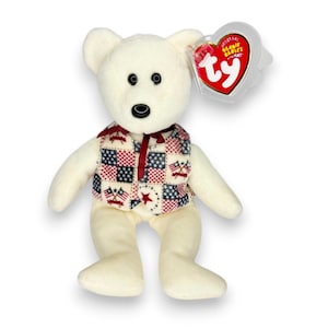 May include: A cream-colored plush bear wearing a vest with a red, white, and blue patchwork pattern featuring stars and crossed flags. The bear has black plastic eyes and a black nose. A heart-shaped Ty Beanie Babies tag is attached to the ear.