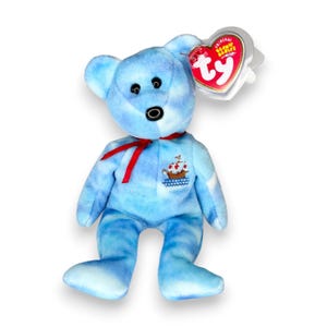 Peut inclure: Un ours en peluche bleu clair avec un motif tie-dye, des yeux et un nez en plastique noir, portant un ruban rouge autour du cou. Un motif de bateau brodé est sur la poitrine. Une étiquette Ty Beanie Babies rouge et blanche est attachée.
