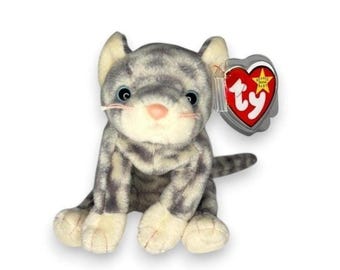 May include: A gray and white tabby cat plush toy with dark gray stripes, a light pink nose, and black eyes. A heart-shaped red and white tag with the letters ty and a yellow star is attached to the ear of the stuffed animal.