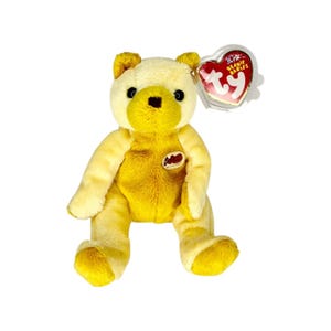 May include: A plush bear toy with light yellow fur on the head and limbs, and a darker golden yellow torso. It features black plastic eyes and a black nose. A heart-shaped tag with the text Beanie Babies and ty is attached to the ear.