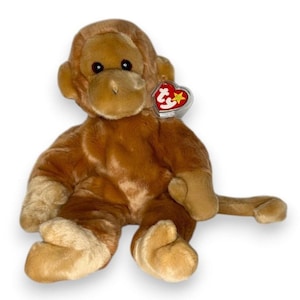 Può includere: Un peluche a forma di scimmia con pelliccia marrone chiaro, tratti del viso marrone scuro e una lunga coda. Il pupazzo ha occhi scuri e un naso piccolo. Un'etichetta Ty rossa e gialla a forma di cuore è attaccata all'orecchio in una custodia.