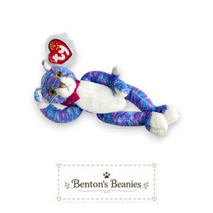 May include: A plush cat toy with a purple and blue tie-dye pattern, a white belly, and white paws. The cat has yellow eyes, a pink nose, and a dark pink bow tie around its neck. A heart-shaped red Ty Beanie Baby tag is attached to the ear.