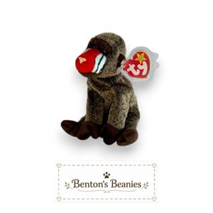 May include: A plush mandrill toy with brown fur, a bright red nose, and white cheek markings. It features a heart-shaped Ty Beanie Baby tag attached to its ear. Below the toy, a rectangular label displays the text Benton's Beanies.