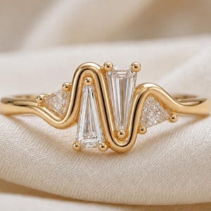 Può includere: Un anello in oro con un design a fascia ondulata, incastonato con due pietre centrali trasparenti a taglio baguette e due pietre trasparenti più piccole a taglio triangolare sui lati. La fascia in metallo ha una finitura lucida e fluida.