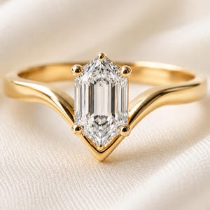May include: A gold ring featuring a single emerald-cut clear gemstone set in a V-shaped prong setting. The band is smooth and polished, with a curved design that tapers toward the center stone, creating a pointed silhouette at the base of the setting.