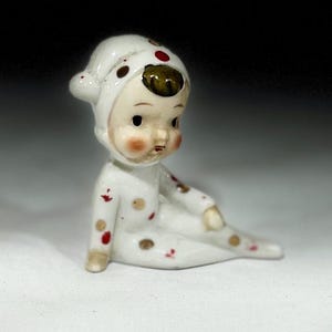 May include: A vintage porcelain figurine of a baby in a white onesie with a hood. The onesie is decorated with red, brown, and gold polka dots. The baby has rosy cheeks and a contemplative expression.