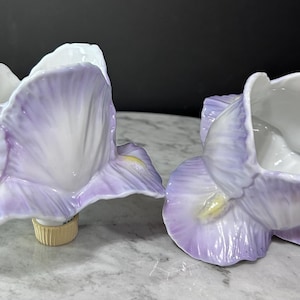 May include: Two ceramic iris flower-shaped vases. The vases are white inside with lavender petals and yellow accents. The vases are on a light gray surface.