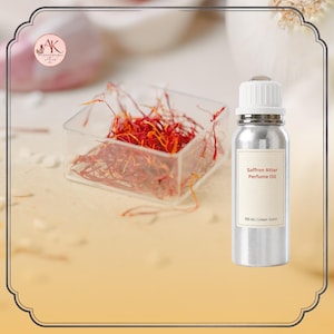 May include: A silver-coloured metal bottle with a white cap and a label reading Saffron Attar Perfume Oil, 100 ml | Unisex Scent. Next to the bottle is a clear square container filled with red and orange saffron threads. The logo AK International Attar is visible.