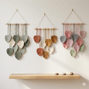May include: Three macrame wall hangings featuring leaf-shaped fibre art suspended from wooden dowels. The left piece uses shades of sage and cream, the centre uses ochre and terracotta, and the right uses dusty pink, grey, and sage green tones.