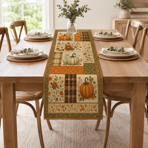 May include: A rectangular table runner featuring a patchwork design of pumpkins, autumn flowers, and plaid patterns in shades of orange, green, and brown. The fabric runner is centered on a wooden table with white plates and woven placemats.