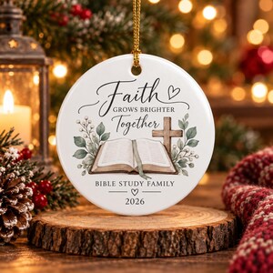 May include: A round white ceramic ornament featuring a gold hanging string. The design includes an open Bible, a wooden cross, and green foliage. Text reads: Faith GROWS BRIGHTER Together, BIBLE STUDY FAMILY, and 2026, with small heart icons.