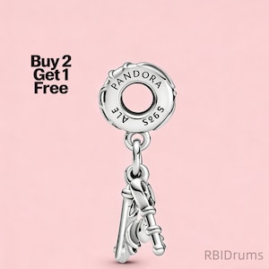 Op de afbeelding: Een zilveren bedel met een camera-ontwerp, hangend aan een ronde Pandora-bedel met de tekst "PANDORA ALE S925". De bedel staat op een roze achtergrond. De tekst "Buy 2 Get 1 Free" staat linksboven.