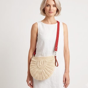 May include: A semi-circular beige crochet handbag featuring a textured fan-like pattern and a vibrant red fabric shoulder strap attached with silver-toned metal hardware. The bag has a structured, woven appearance with a distinct central arch design.