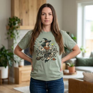 Può includere: Una t-shirt a maniche corte color verde salvia con un'illustrazione grafica di una strega con cappello, che tiene in mano un cocktail e cavalca una scopa. Il testo "Practical Magic" è stampato in un carattere stilizzato, accompagnato da pipistrelli.