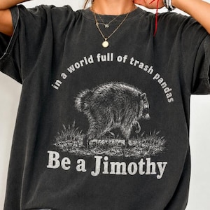 Puede incluir: Una camiseta gris carbón de manga corta con un gráfico de un mapache de pie sobre la hierba. El texto sobre el mapache dice "in a world full of trash pandas" y el texto debajo dice "Be a Jimothy" en una fuente blanca desgastada.
