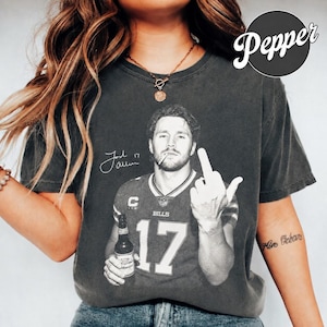 May include: A charcoal gray short-sleeve t-shirt featuring a black and white graphic of a football player in a jersey with the number 17, holding a beer bottle and giving a middle finger gesture. The graphic includes a signature and the text Bills.