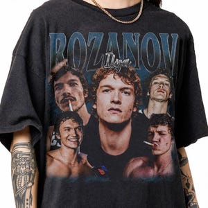 May include: A charcoal grey short-sleeve t-shirt featuring a graphic print of five different portraits of the same person. The text ROZANOV is printed in large blue letters at the top, with the name Ilya written in a smaller, stylised white font below.