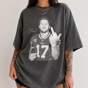 May include: A charcoal gray oversized t-shirt featuring a black and white graphic of a football player in a jersey with the number 17, holding a beer bottle, smoking a cigarette, and gesturing with a middle finger, with a signature and the text Josh Allen.
