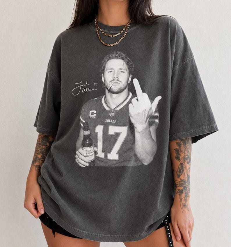 May include: A charcoal gray oversized t-shirt featuring a black and white graphic of a football player in a jersey with the number 17, holding a beer bottle, smoking a cigarette, and gesturing with a middle finger, with a signature and the text Josh Allen.