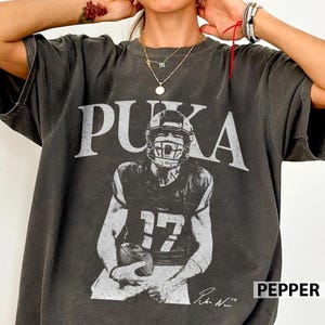 May include: A charcoal grey short-sleeve t-shirt featuring a distressed white graphic of a football player wearing a jersey with the number 17. The text PUKA is printed in large white capital letters above the player, with a signature below.