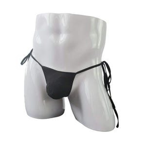 May include: A black thong with thin side ties, featuring a minimalist design with a contoured front pouch. The garment is displayed on a white mannequin torso, highlighting the simple construction and the adjustable tie-side closure mechanism.
