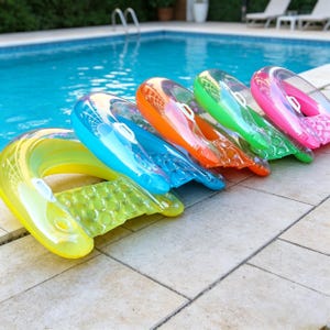 Puede incluir: Una fila de flotadores inflables de piscina de colores amarillo, azul, naranja, verde y rosa. Cada flotador tiene un respaldo curvo transparente y un asiento con un patrón texturizado. Los flotadores están colocados sobre una superficie embaldosada junto a una piscina.