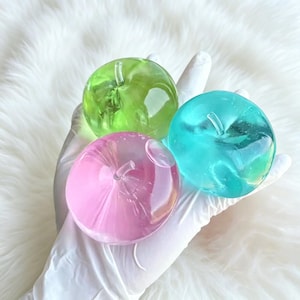 May include: Three translucent apple-shaped ornaments in shades of green, pink, and teal. Each has a stem and a smooth, glossy surface. The ornaments are held in a gloved hand against a white, fluffy background.